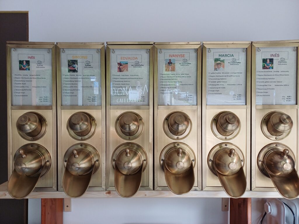 an der Wand hängen goldene Kaffeebehälter mit Kurzvorstellungen der jeweiligen Produzentin.